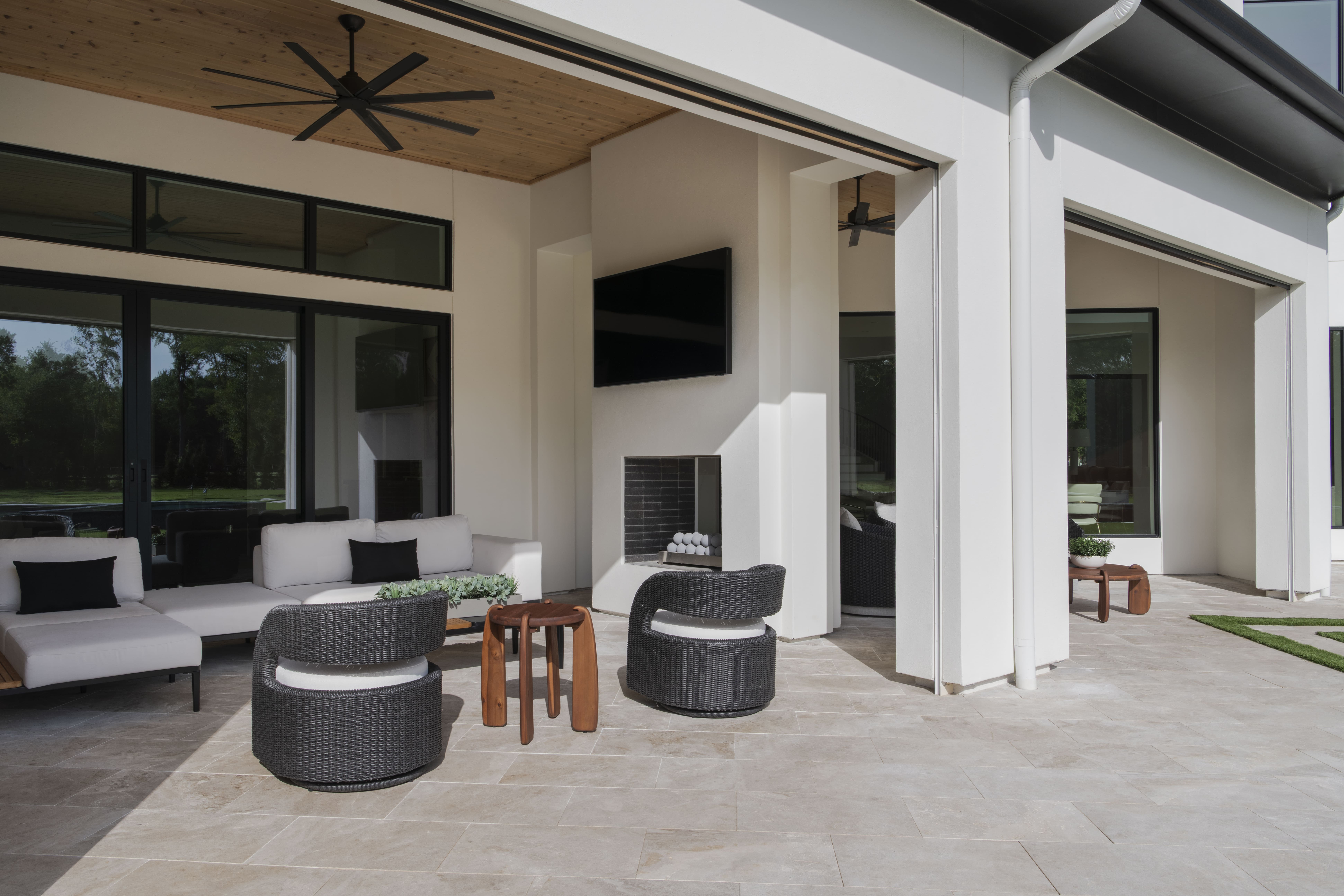 Covered outdoor lounge with modern wicker seating, wood plank ceiling, large black fan, and a fireplace with a mounted TV.