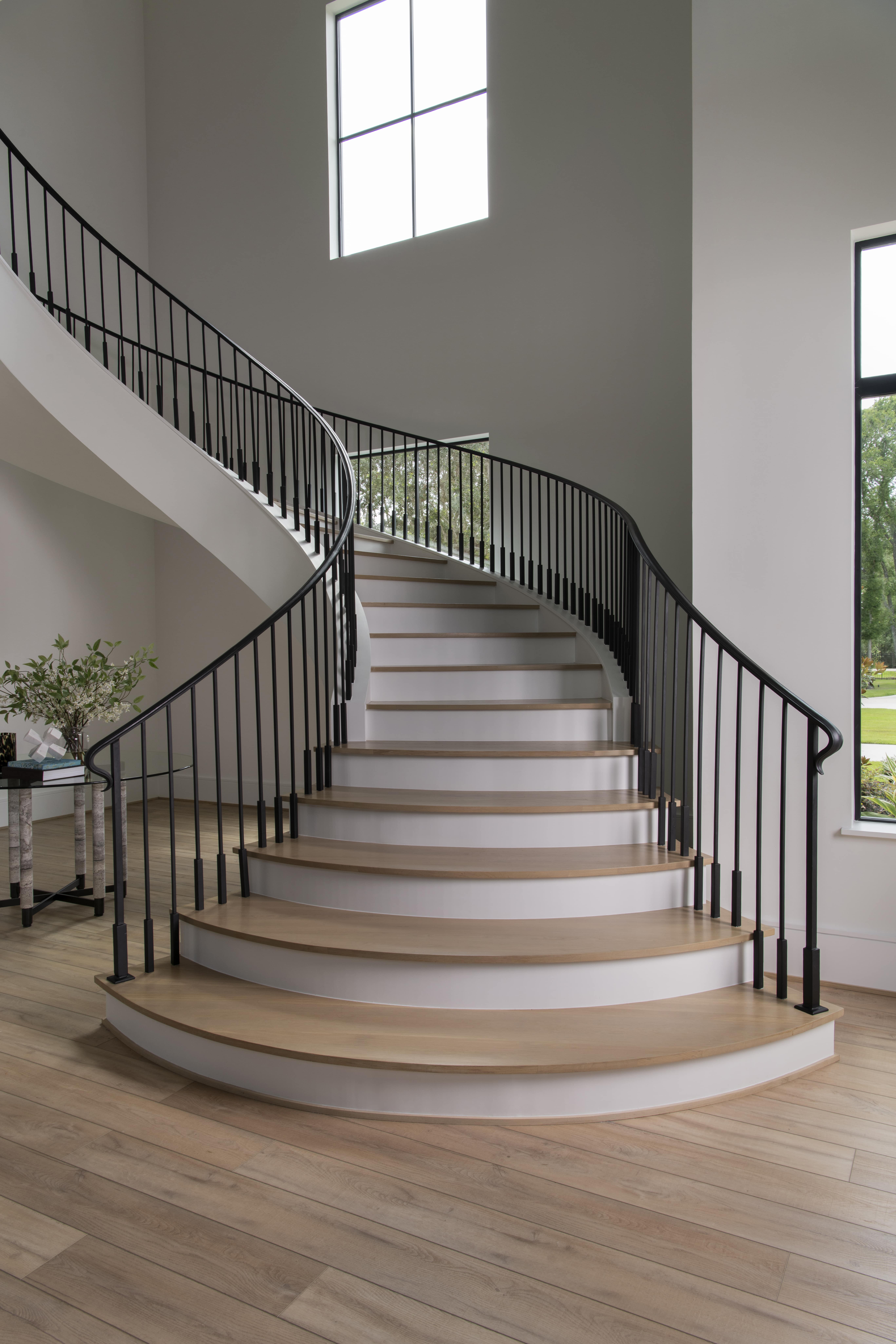 Grand curved staircase with black railings, wood treads, and tall windows filling the space with natural light.