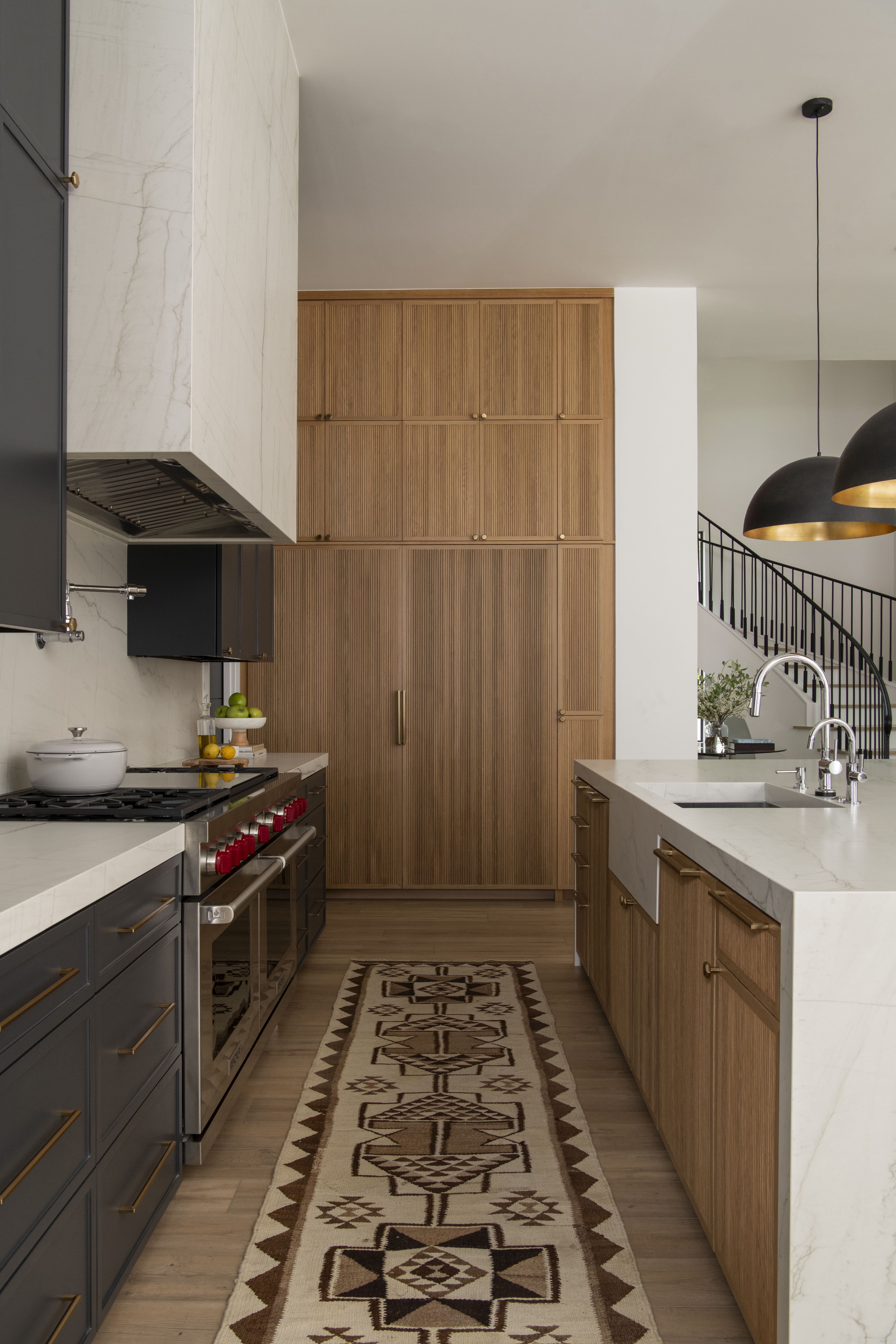 Kitchen range wall with dark cabinetry, marble hood, integrated double ovens, wood-paneled refrigeration, and a patterned area runner.