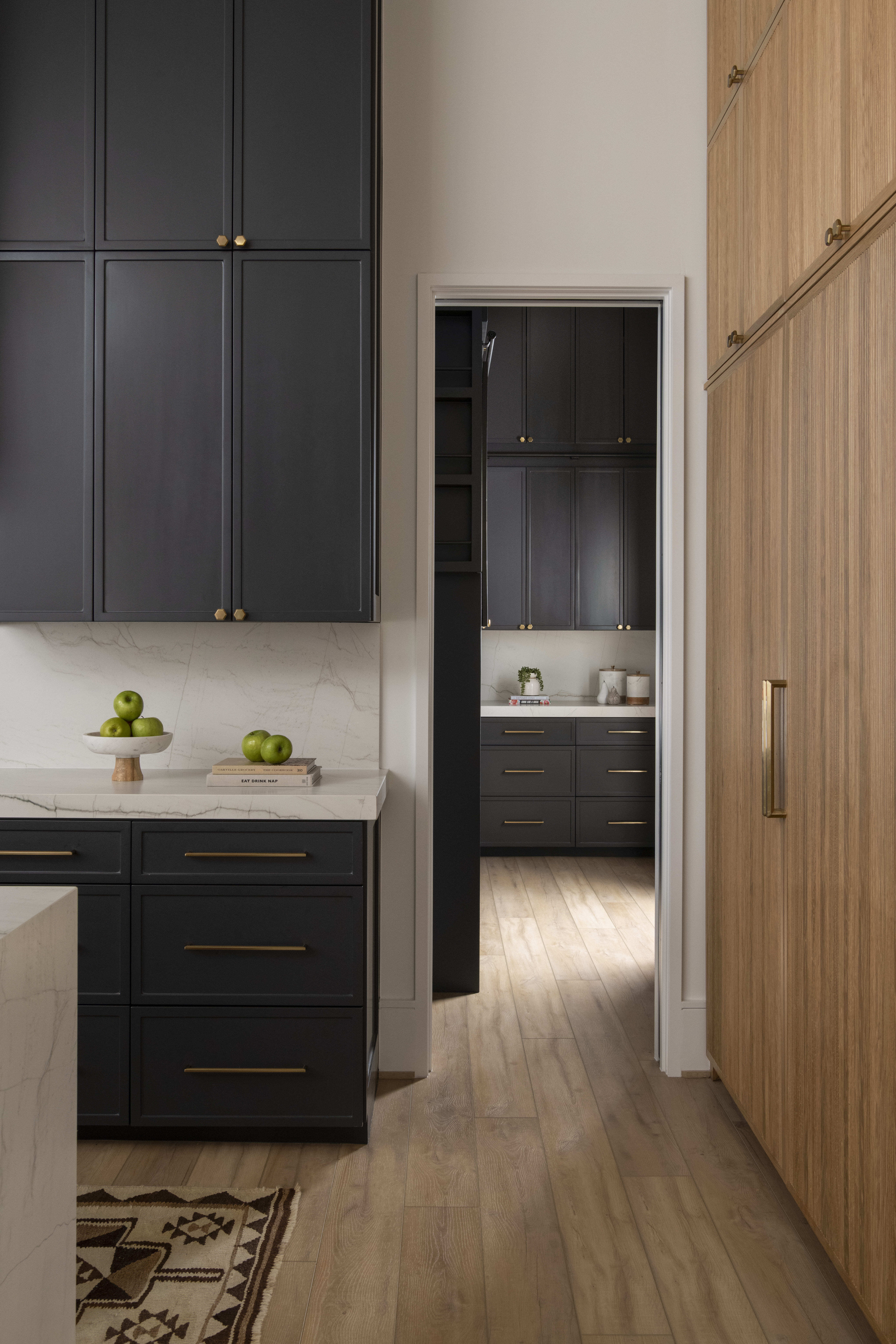 Two-room pantry with dark cabinetry, marble counters, and wood flooring leading into a secondary prep area.
