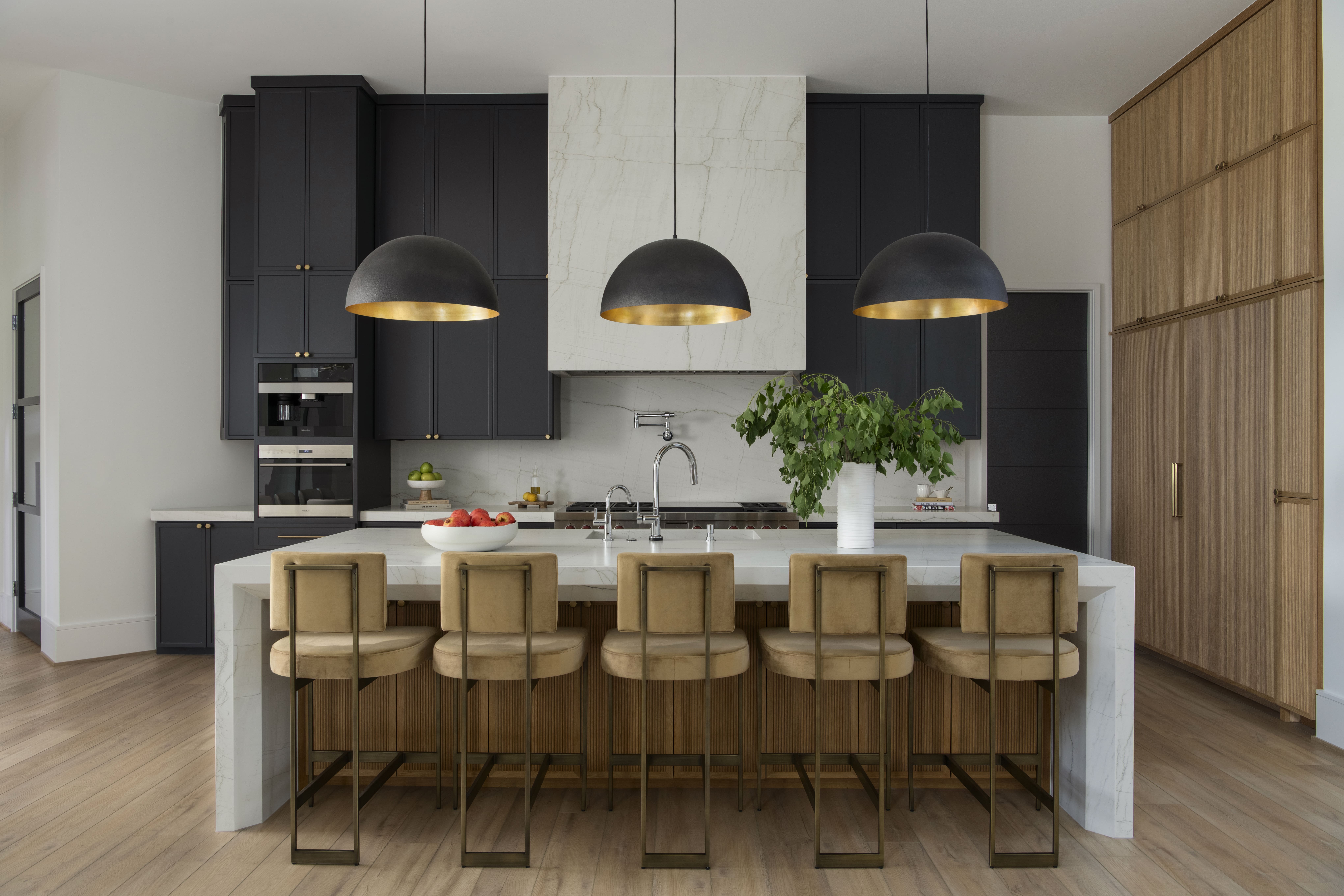 Contemporary kitchen with dark cabinetry, marble hood surround, brass-lined pendant lights, and a large island with upholstered barstools.