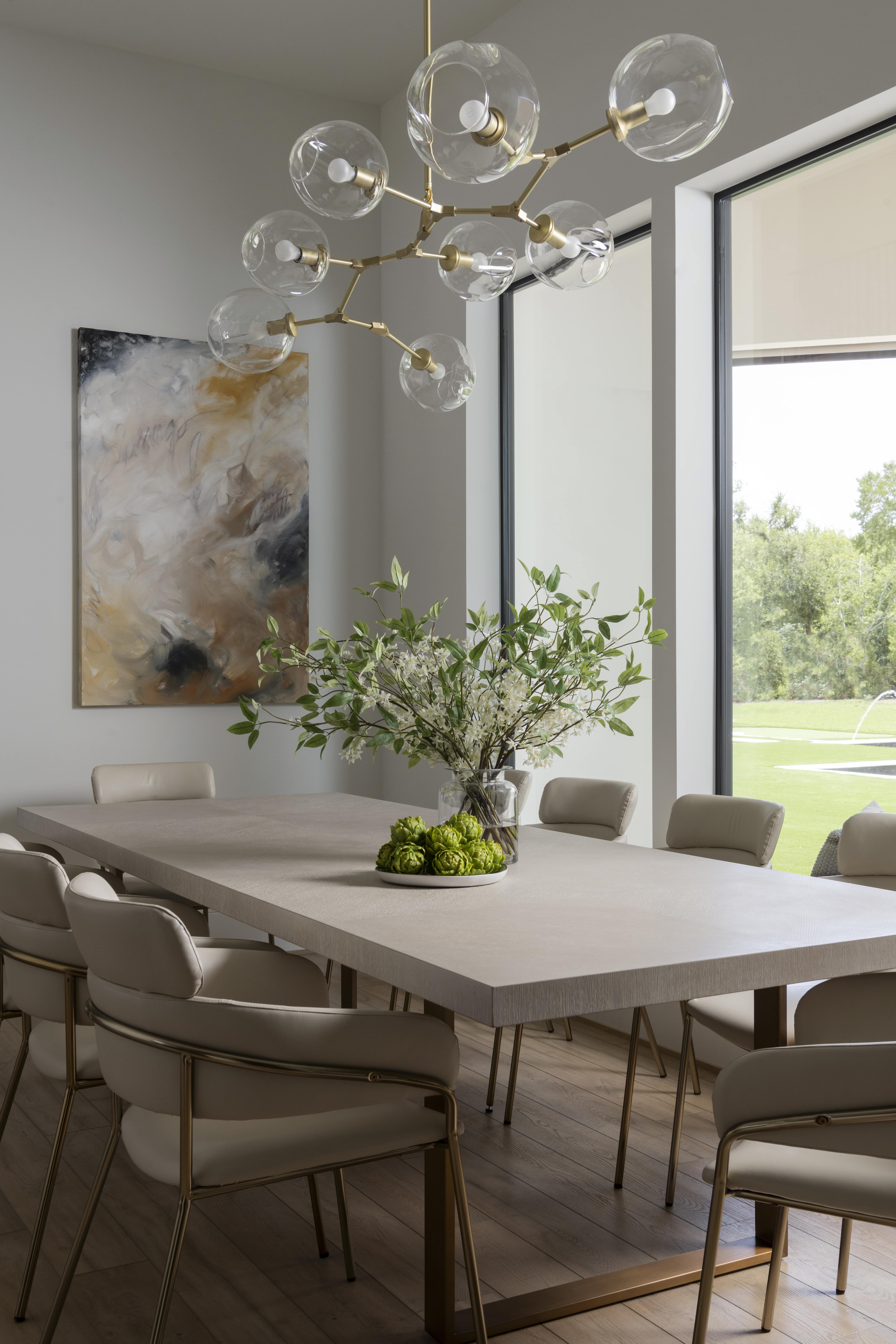 Contemporary dining room with a light wood table, sculptural brass chandelier, and large windows overlooking the lawn.