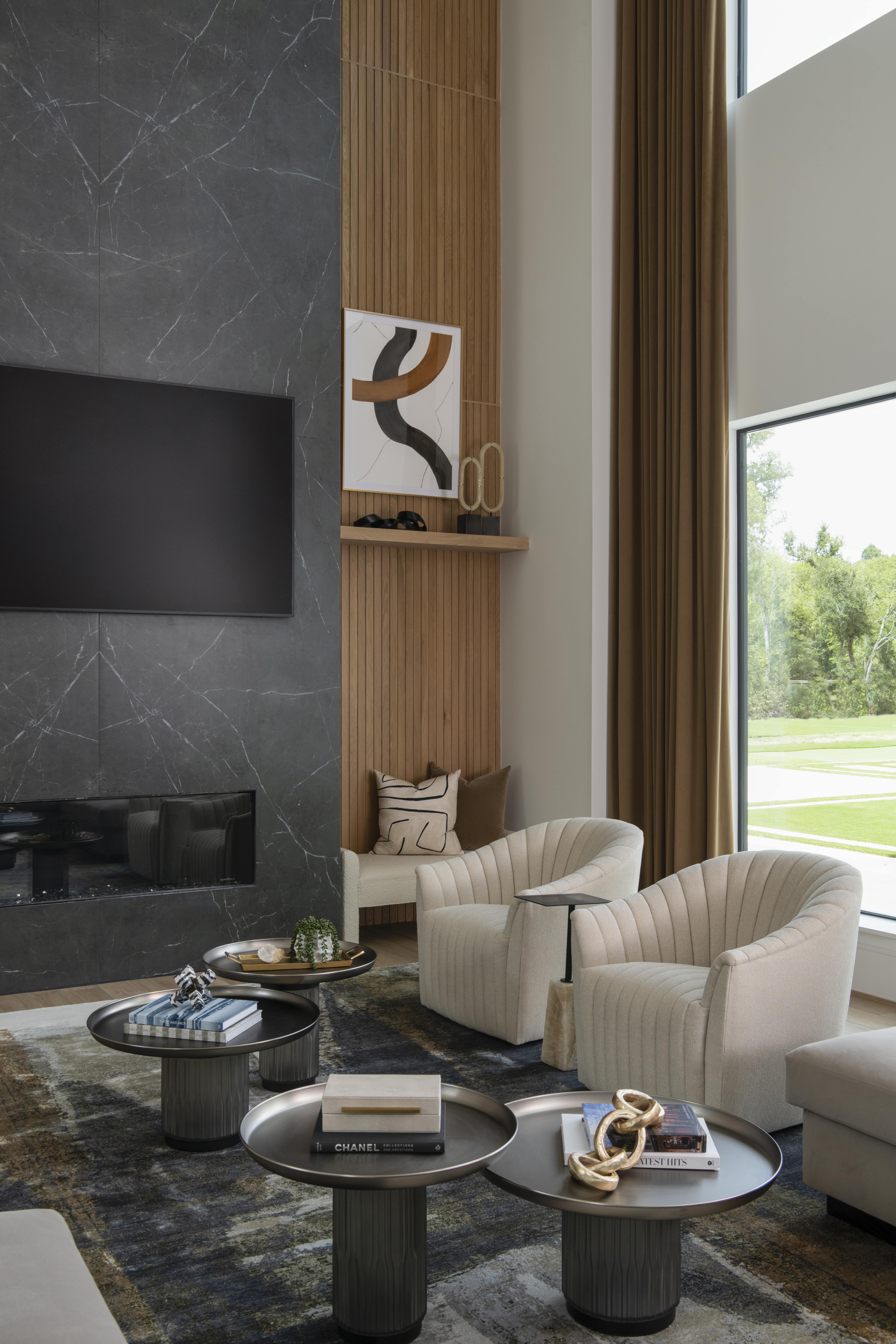 Modern living room corner featuring a floor-to-ceiling dark stone fireplace, vertical wood paneling, soft upholstered swivel chairs, and layered coffee tables styled with books and decor.