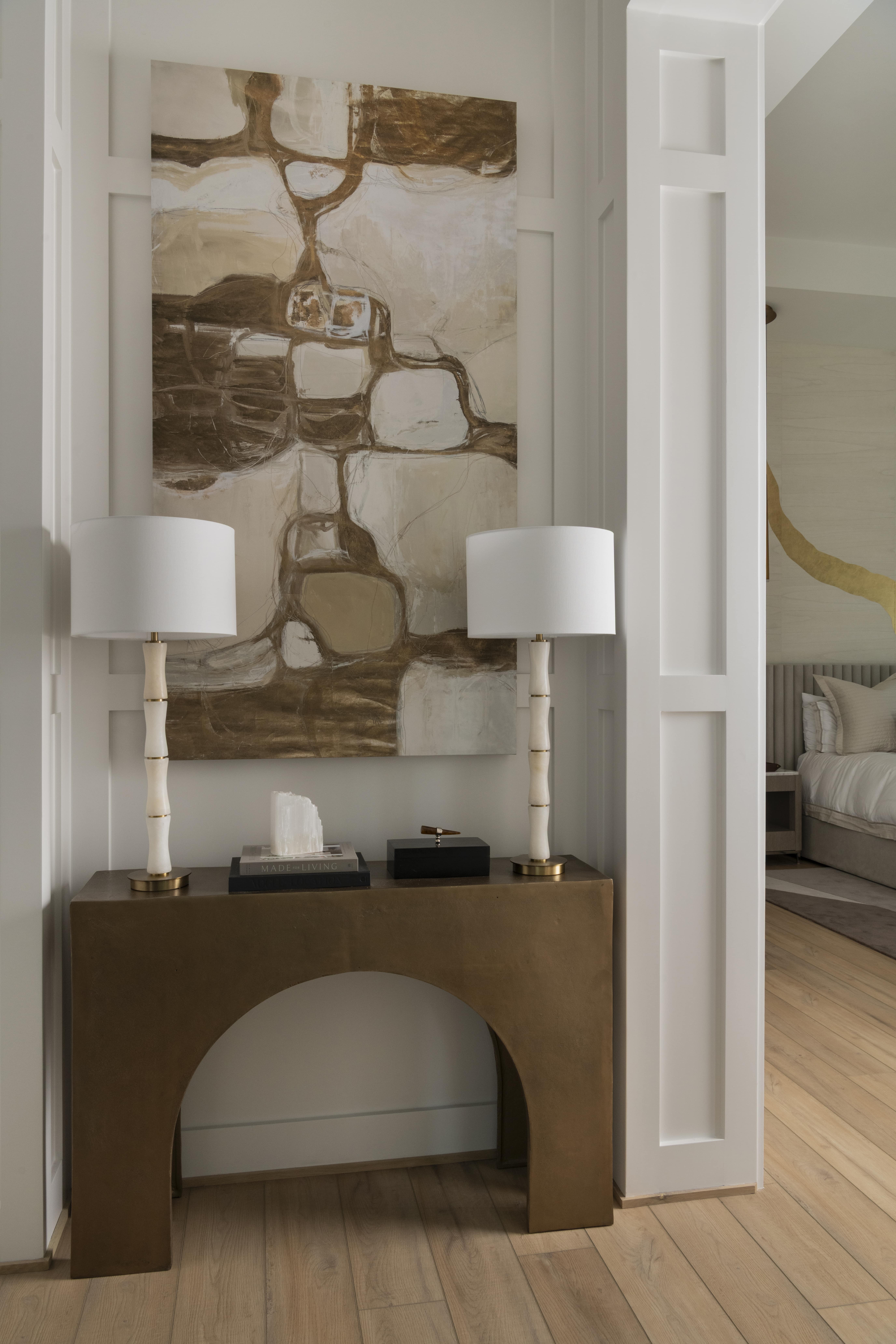 Entry vignette with a sculptural brass console table, abstract artwork, and matching white lamps.