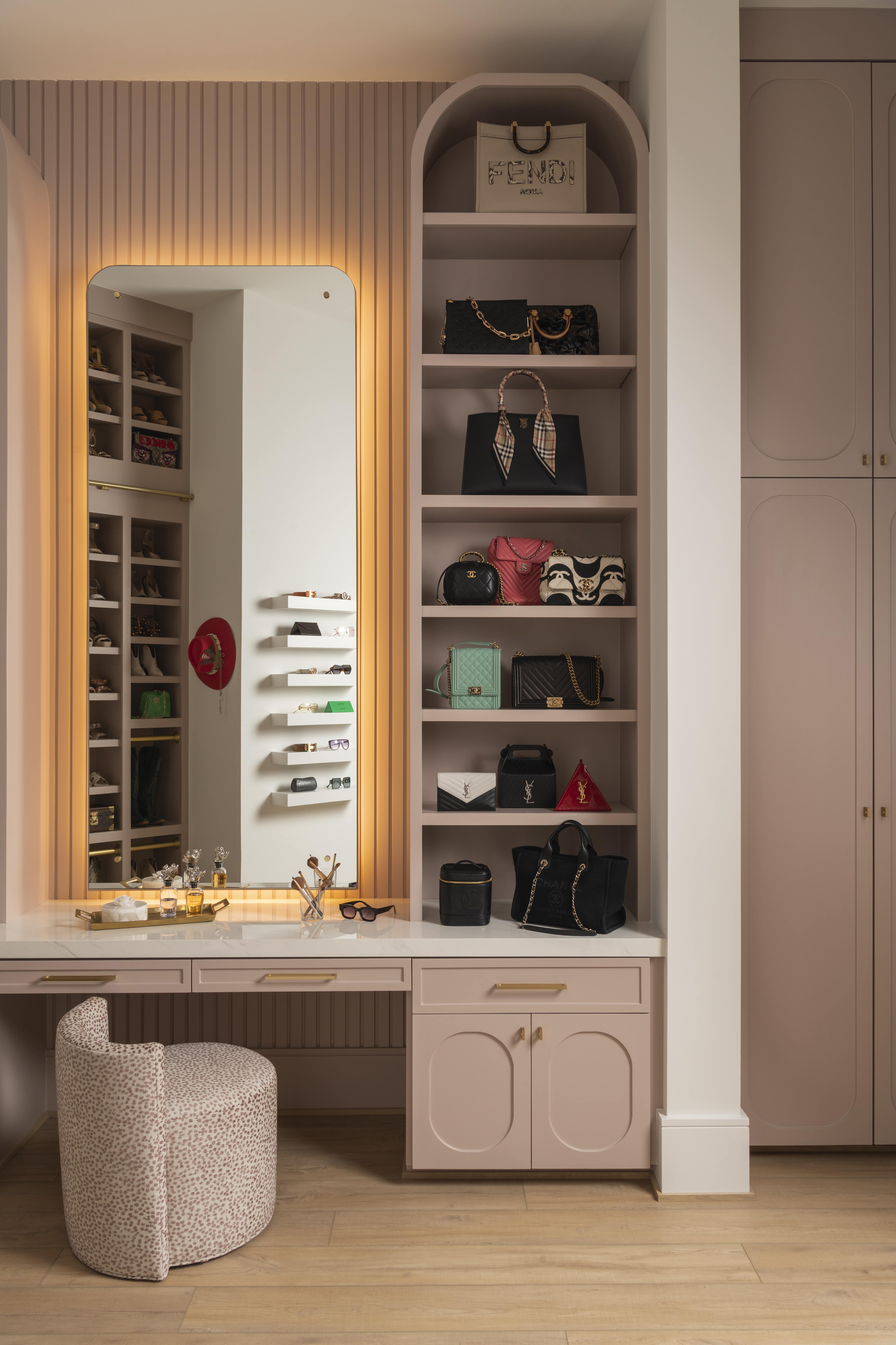 Elegant vanity and shelving display in a blush-toned dressing room, featuring a backlit mirror, curated designer handbags, and built-in storage with brass accents.