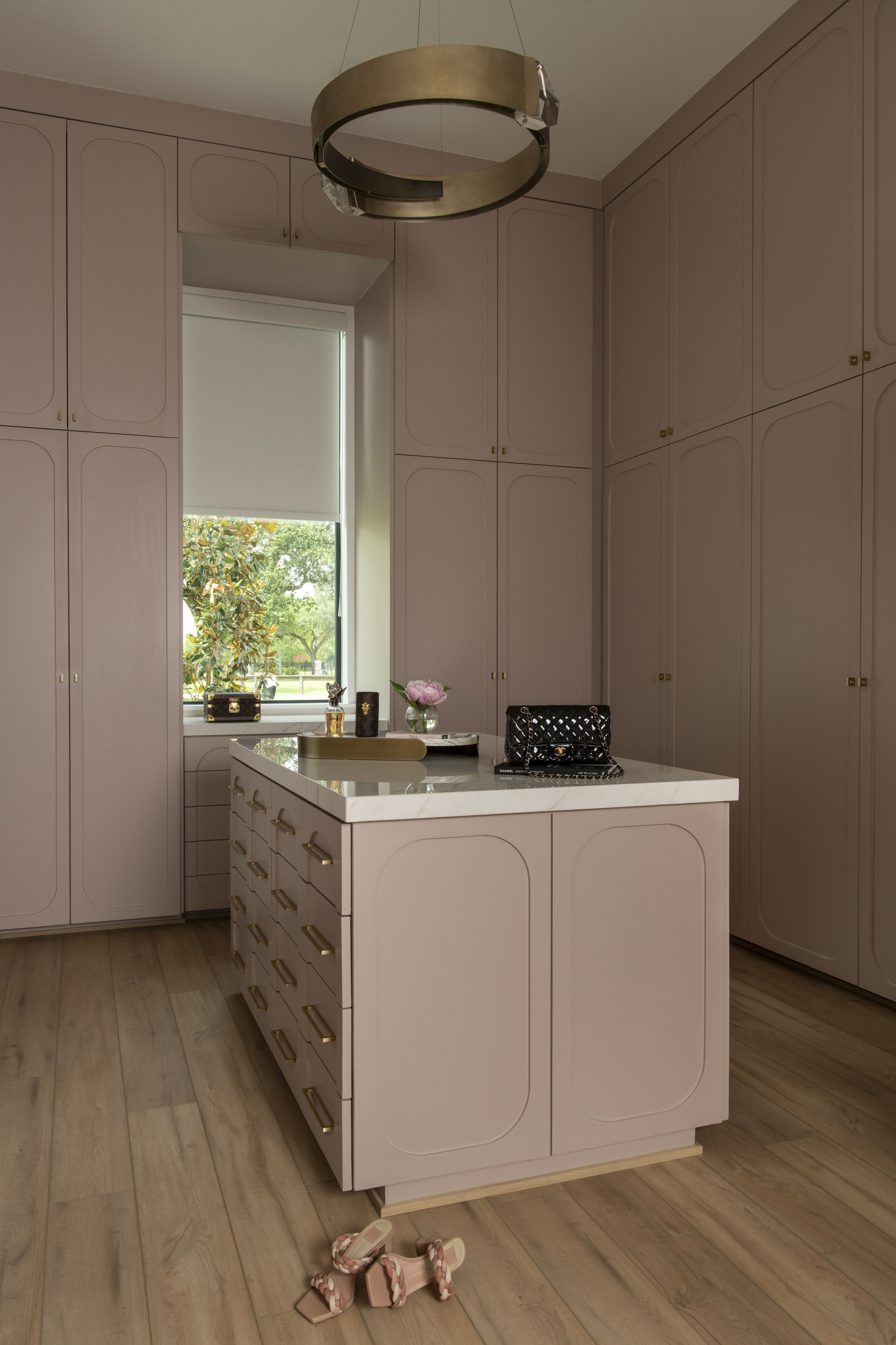 Blush dressing room with full-height cabinetry, brass hardware, a marble-top island, and a sculptural ring chandelier.