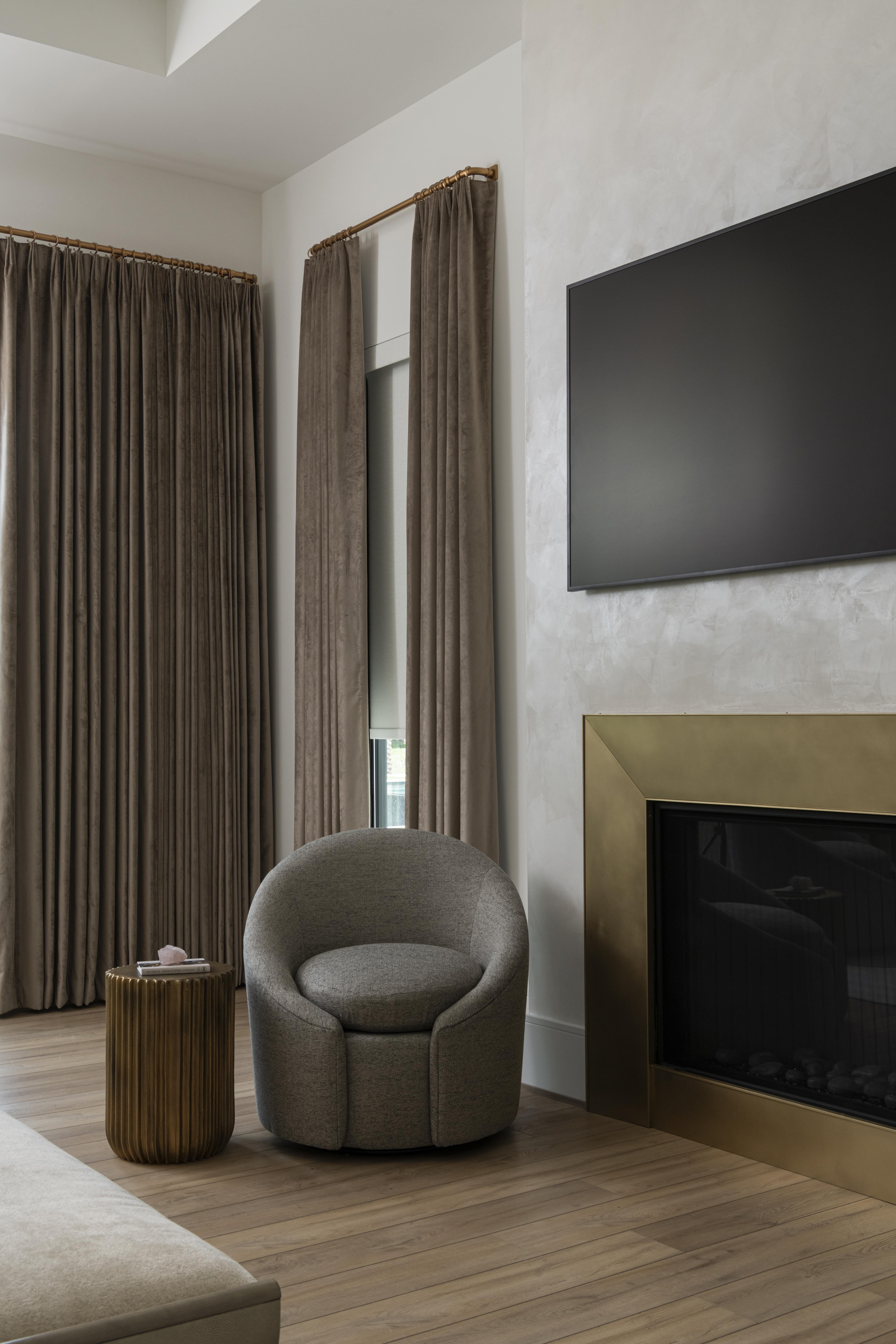 Cozy sitting area with tall taupe drapery, a textured round chair, and a fireplace framed in brushed brass beneath a mounted TV.