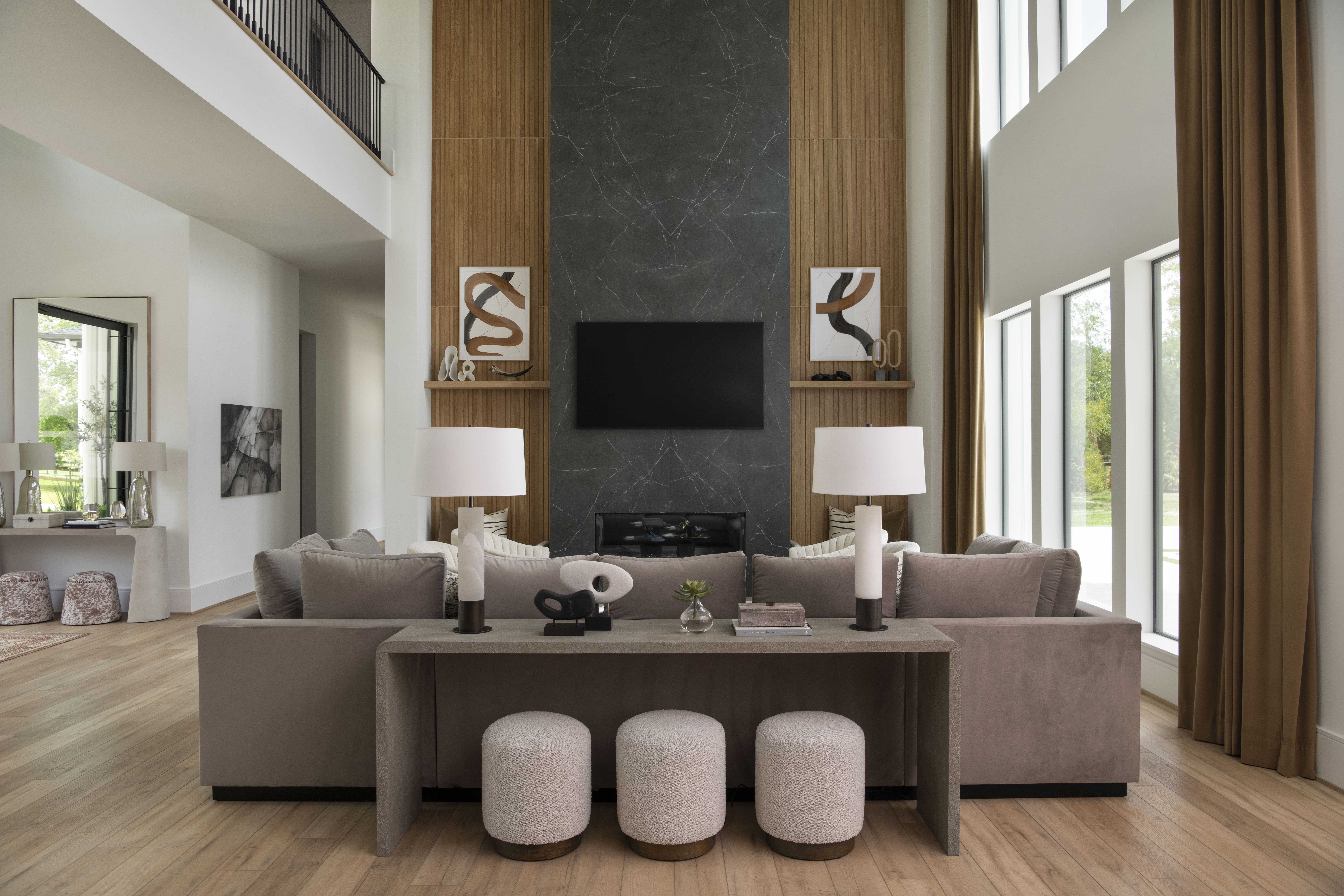 Living room with double-height windows, a dark stone fireplace, wood panel accents, and neutral modern furniture.