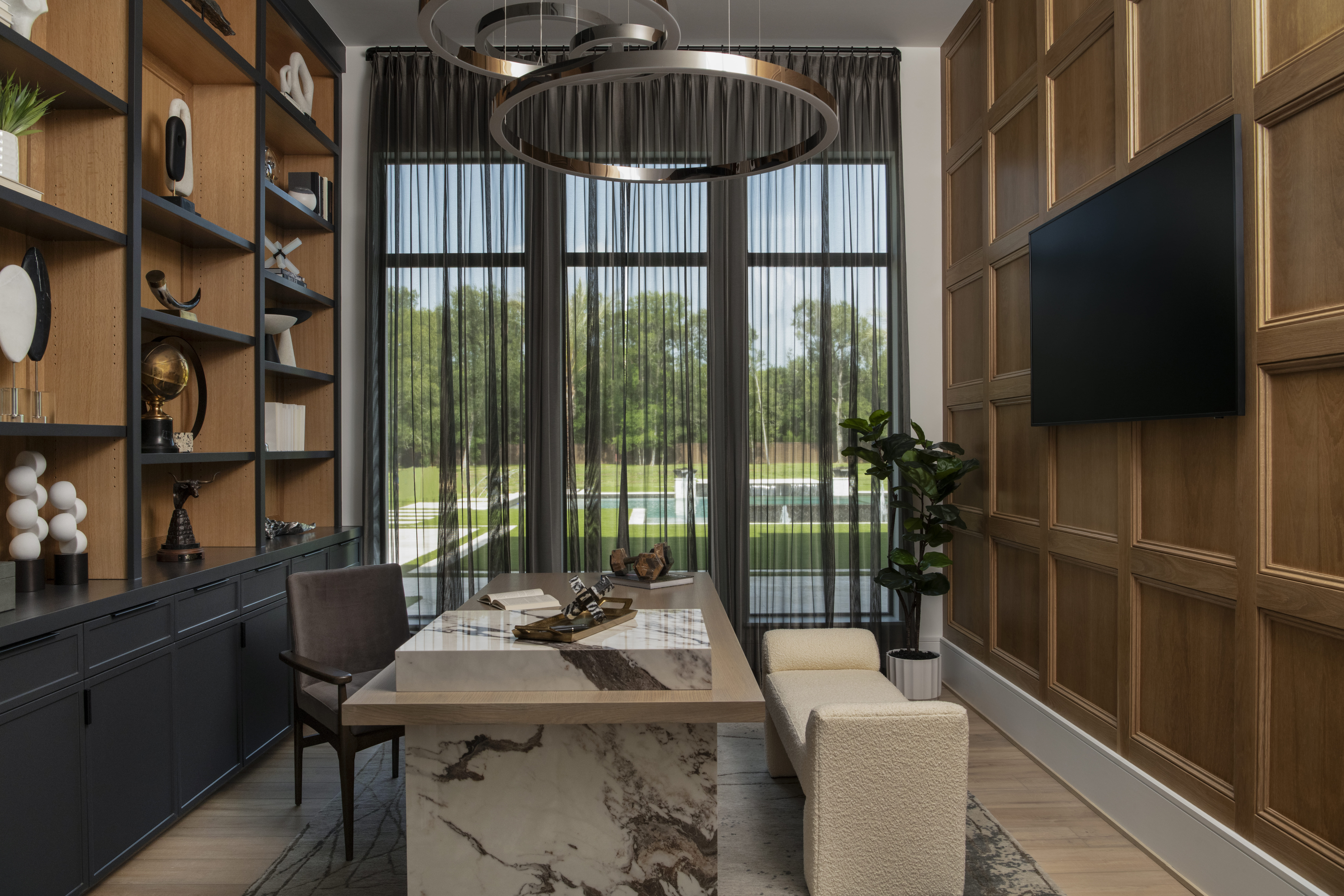 Home office with floor-to-ceiling built-ins, a marble desk, wood paneling, and large windows with sheer drapery overlooking the backyard.