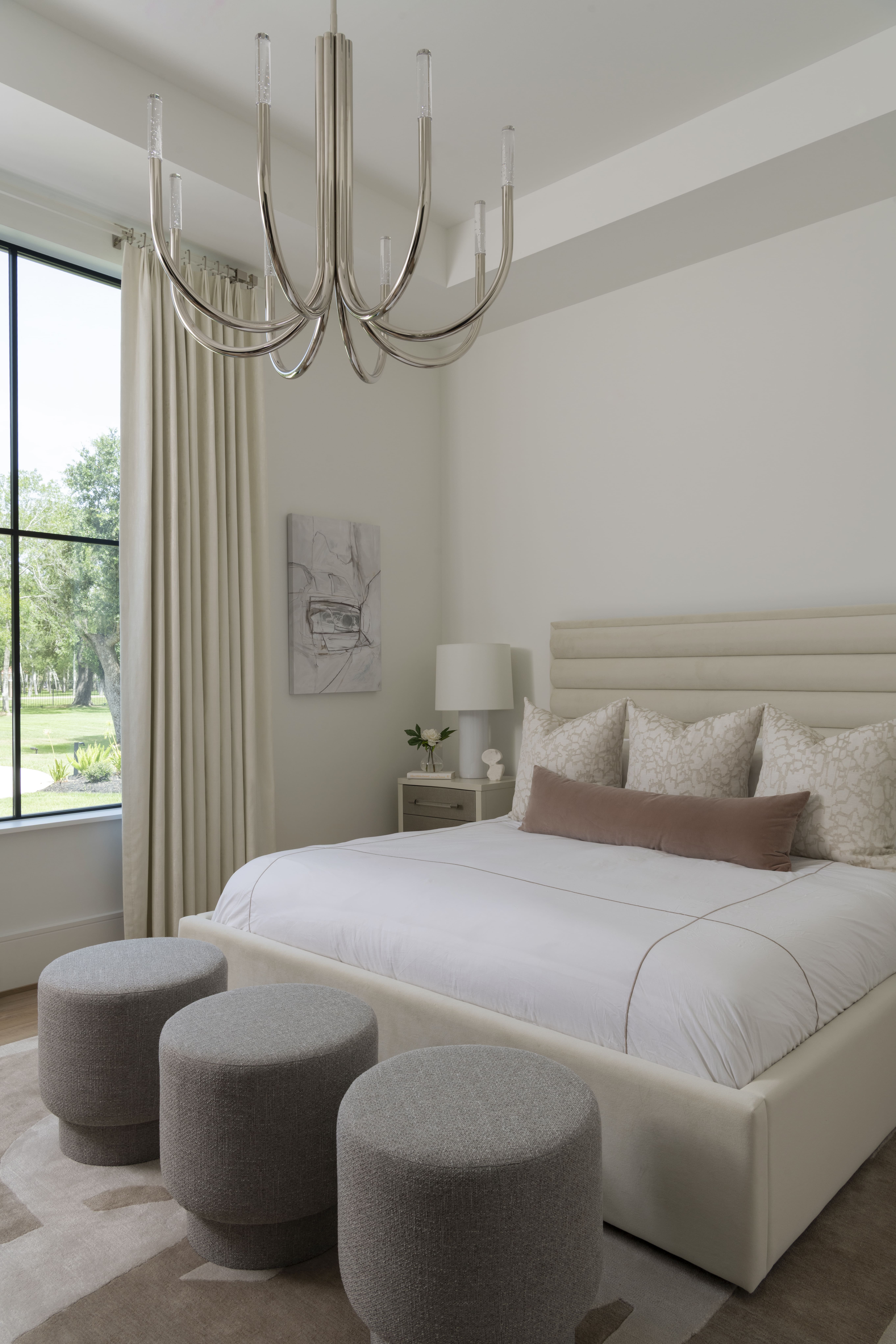 Serene bedroom with upholstered bed, layered neutral bedding, three gray ottomans, and polished chrome chandelier.