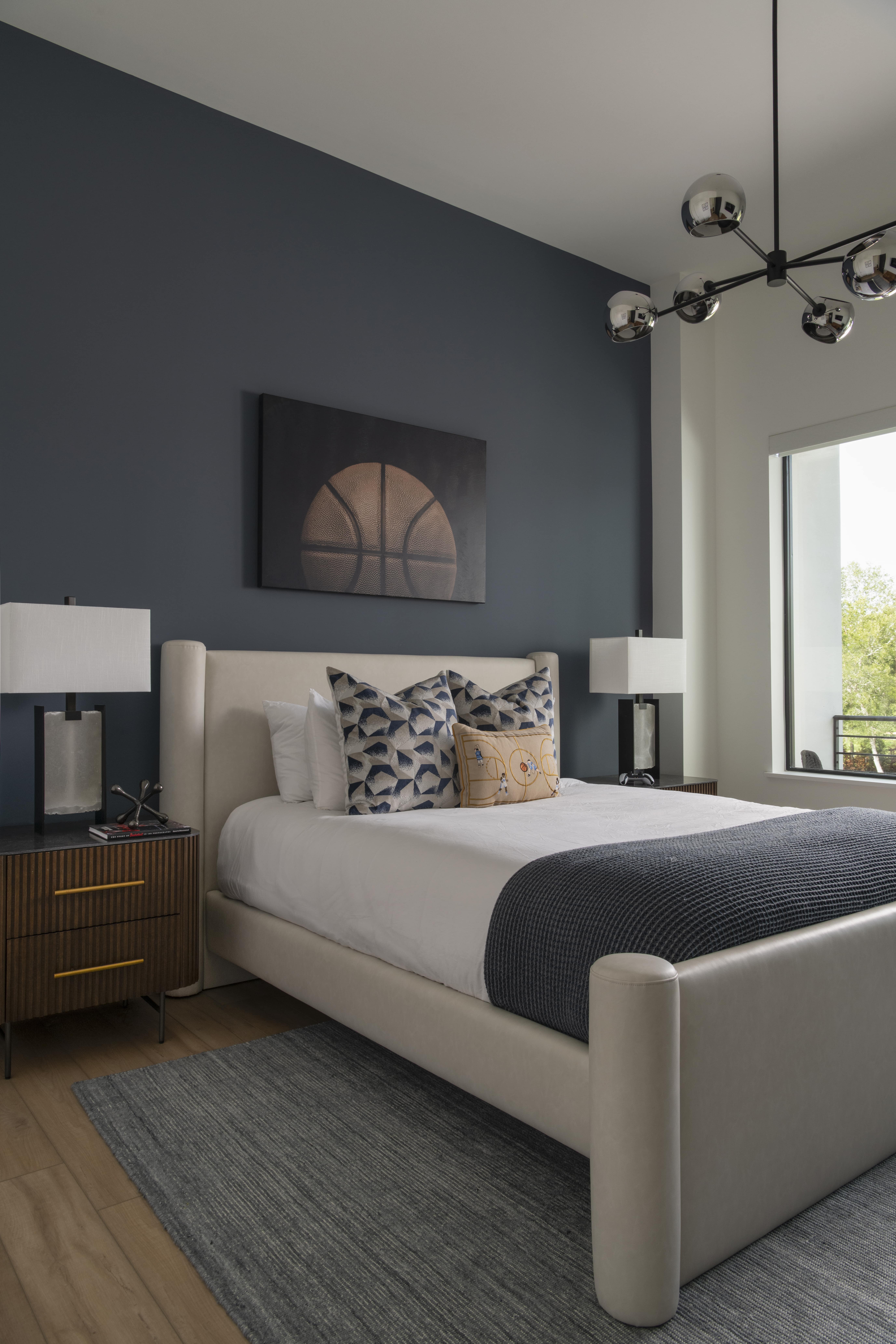 Modern bedroom with navy accent wall, basketball artwork, upholstered bed, and matching nightstands with lamps.