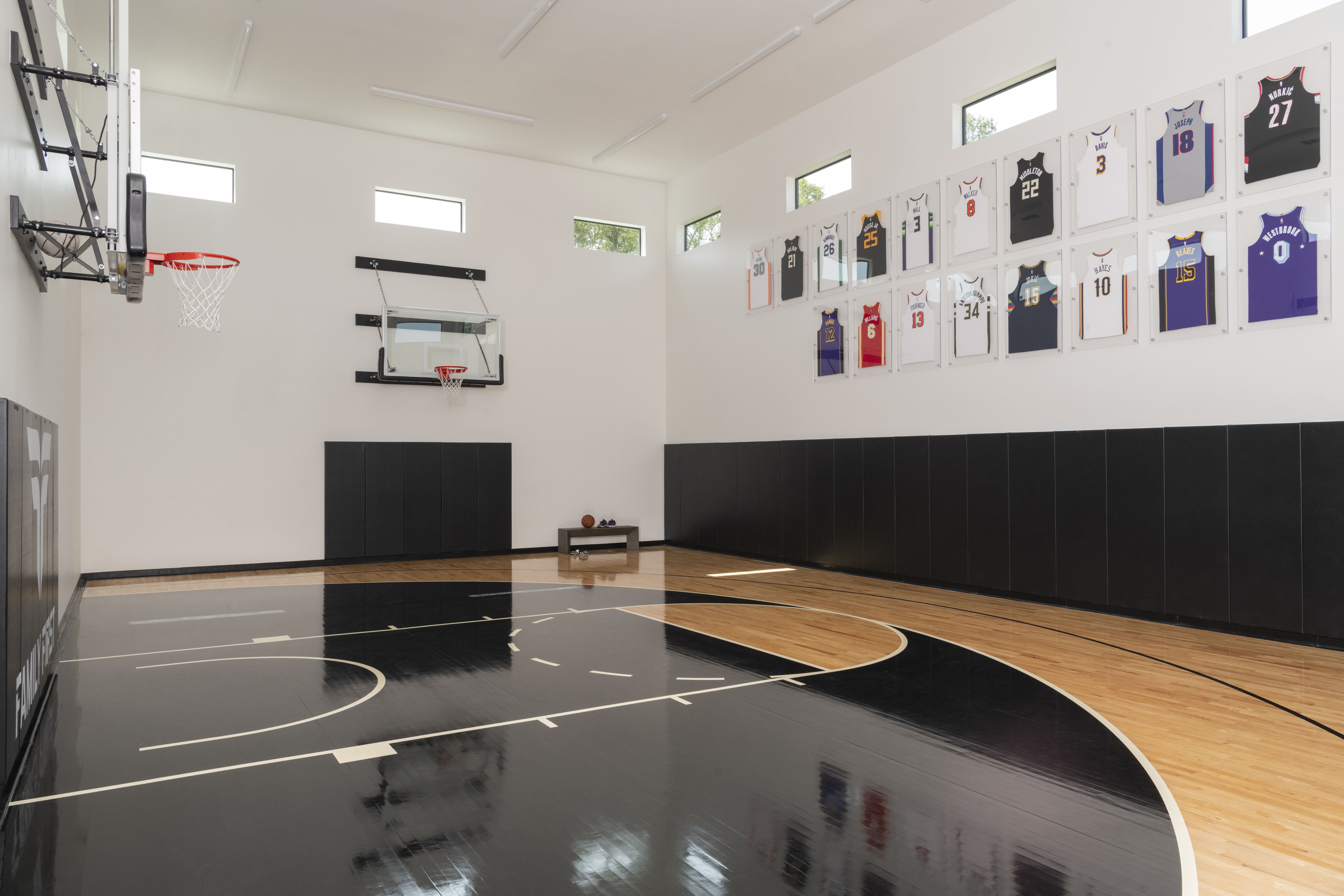 Luxury indoor basketball court with polished black-and-wood flooring, twin wall-mounted hoops, and framed NBA jerseys on display.